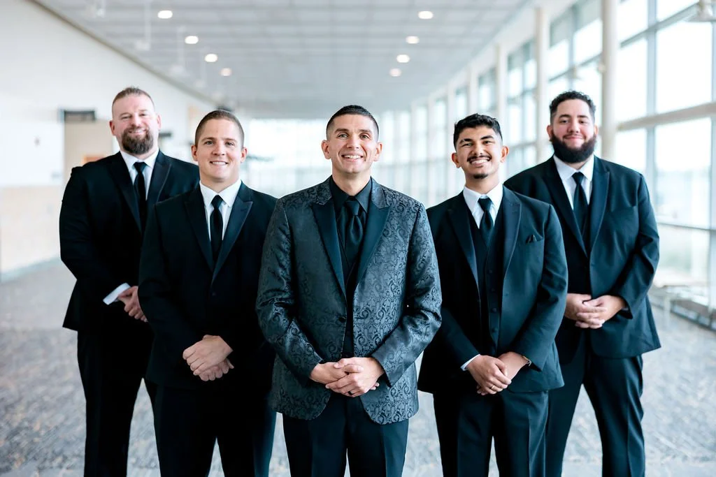 Group of five men in suits standing in a bright indoor corridor.