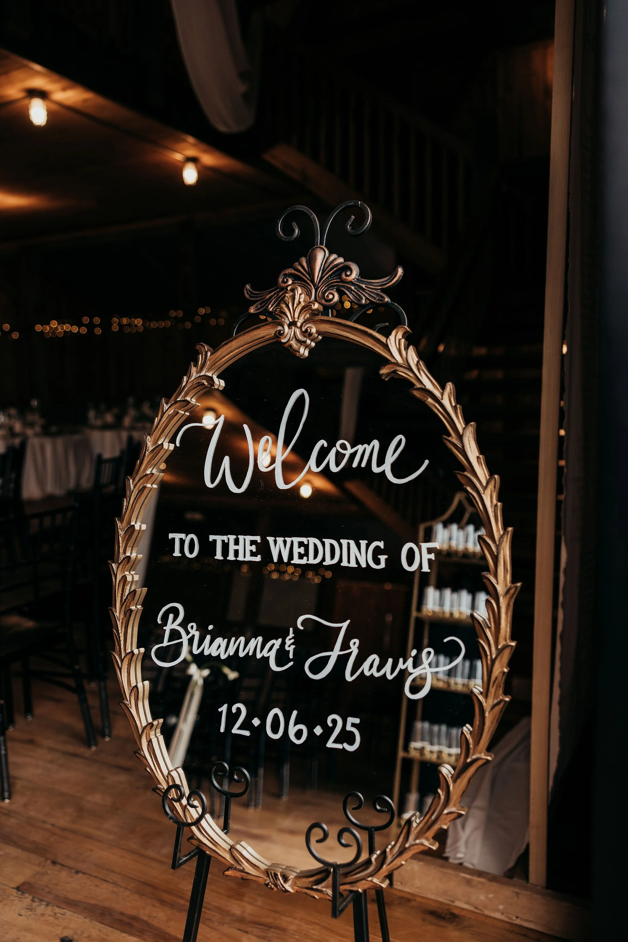 Decorative mirror with the word 'Welcome' and wedding details in white script, ornate gold frame, standing on a black metal easel, in a dimly lit reception space with string lights and tables in the background.