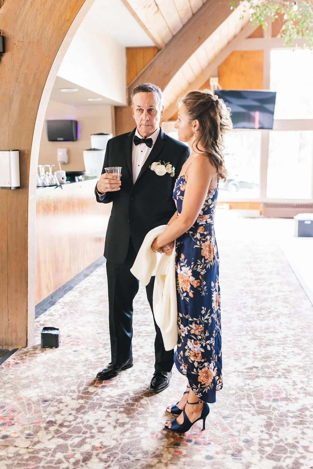 A man in a tuxedo with a boutonniere holding a drink converses with a woman in a floral dress and high heels at an indoor event with wooden beams and large windows.