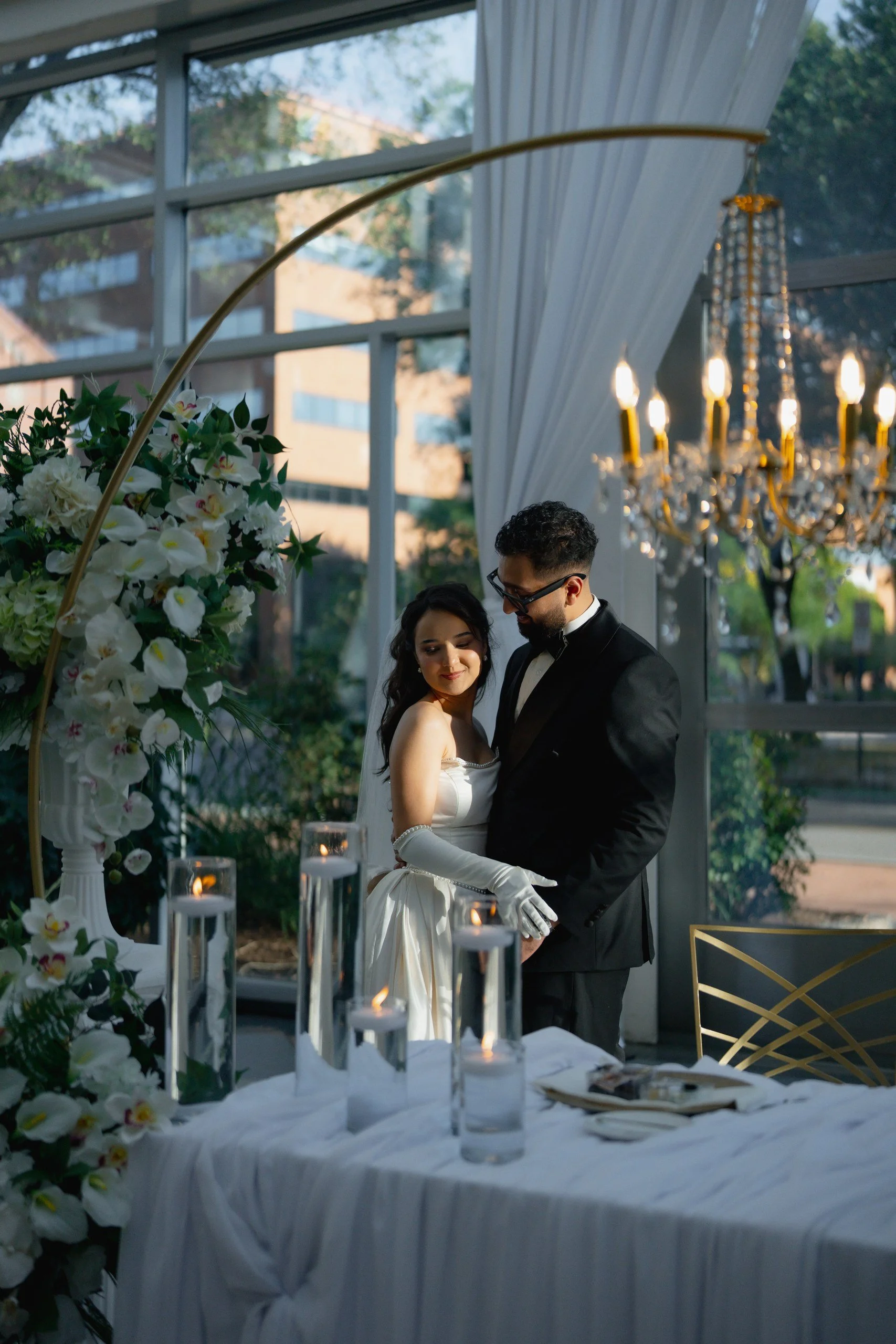 A bride and groom sharing a dance at their wedding reception inside a glass-walled venue, with a floral arrangement and lit candles on a table in the foreground, and elegant chandelier lighting and trees outside.