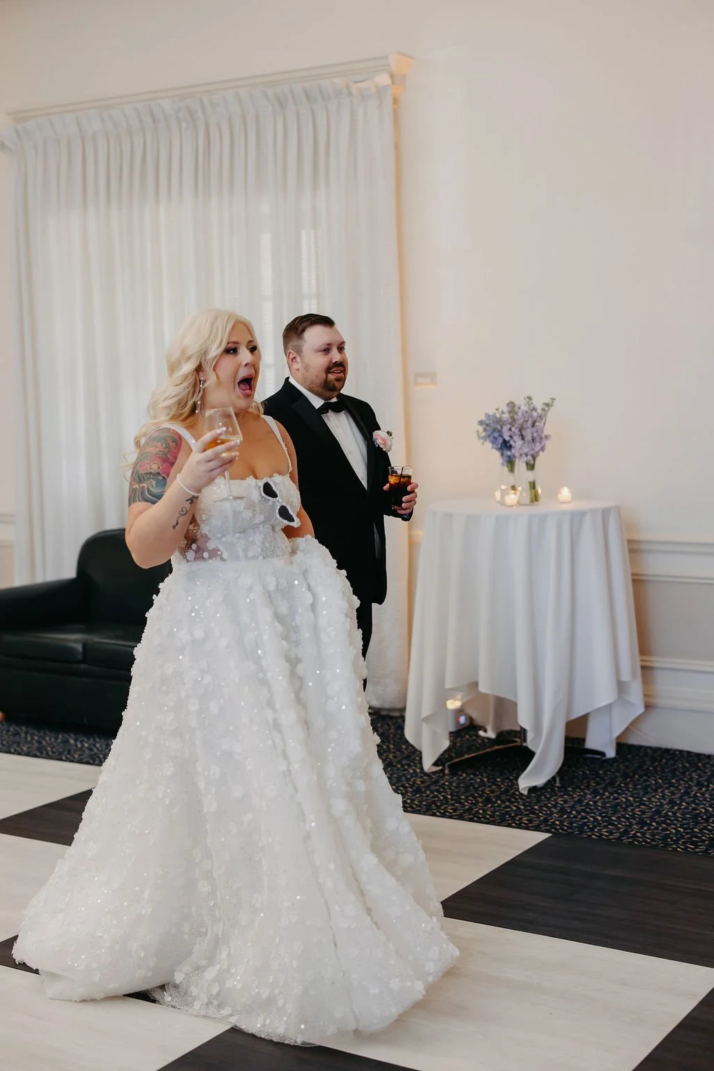 A bride in a white wedding gown with floral details, holding a glass of wine, standing next to a groom in a tuxedo with a boutonniere, in a decorated indoor venue with candles and flowers.
