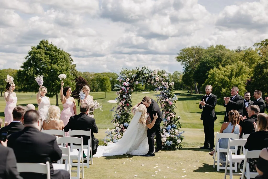 A wedding ceremony outdoors with a bride and groom kissing under a floral arch, surrounded by bridesmaids, groomsmen, and guests on a lush green golf course.