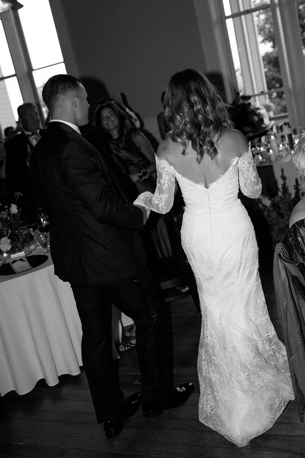 A bride and groom holding hands during their wedding reception, with guests in the background, in a room with large windows.