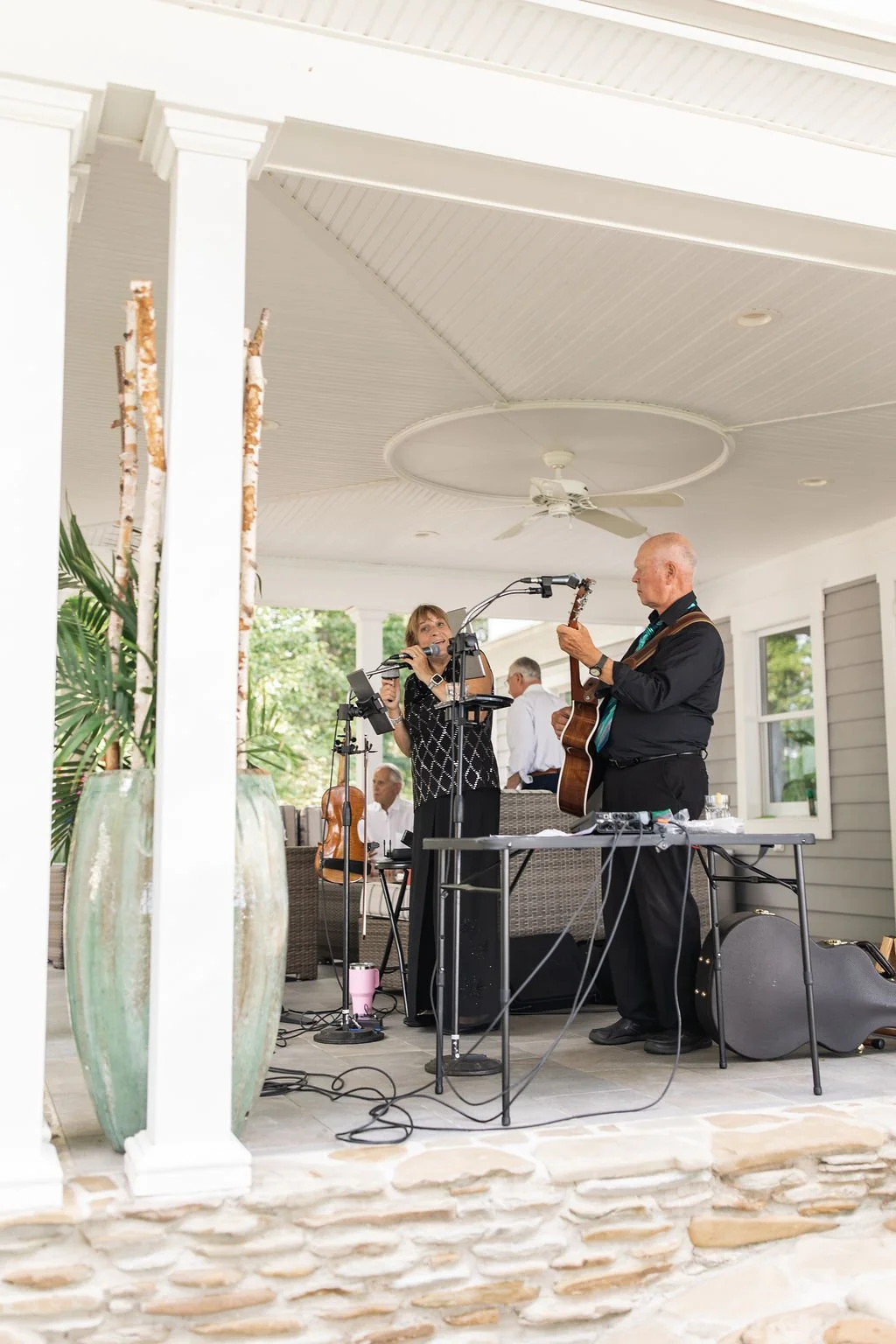 Two musicians performing on a covered porch, one woman singing and playing a tambourine, one man playing guitar, with other people seated in the background.