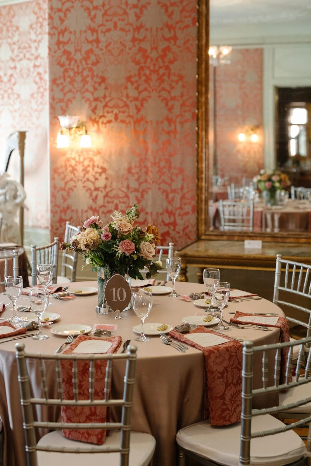Elegant round banquet table set for a formal event with a centerpiece of pink and cream roses, surrounding table settings with pink napkins, wine glasses, and plates, in an opulent room with a pink damask wallpaper, large mirror, and chandelier light