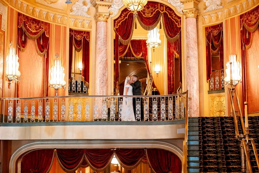 A bride and groom kissing on a balcony inside an elegant, ornate theater or ballroom with red curtains, gold accents, and vintage lighting fixtures.