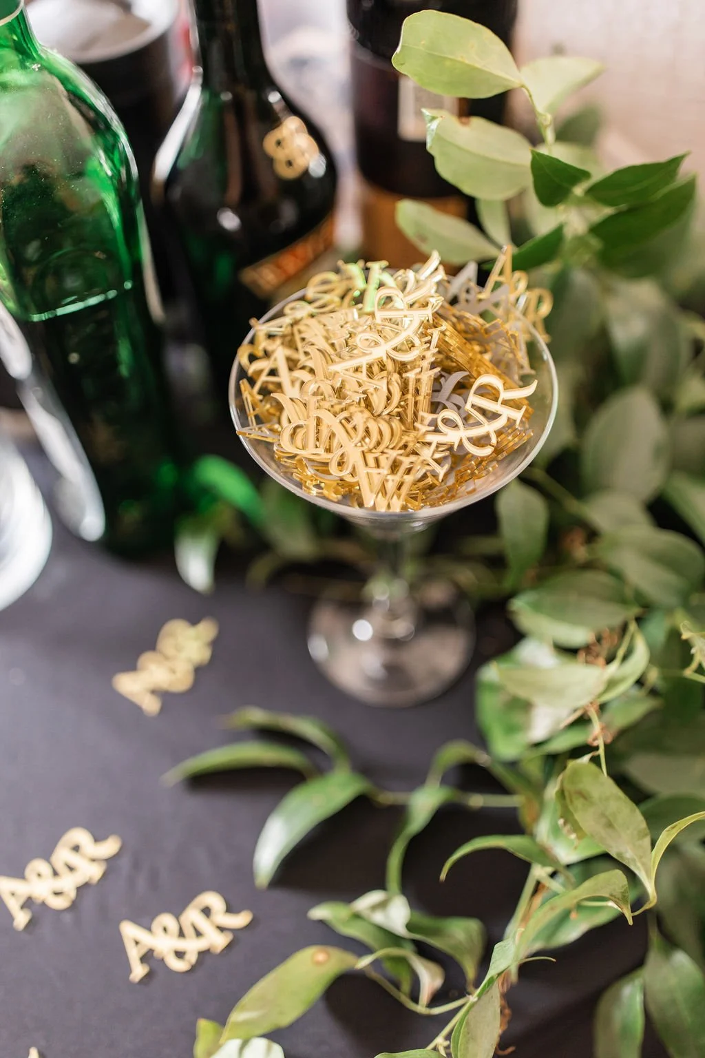 A tall glass filled with golden letter-shaped decorations, surrounded by green leaves and bottles in the background.