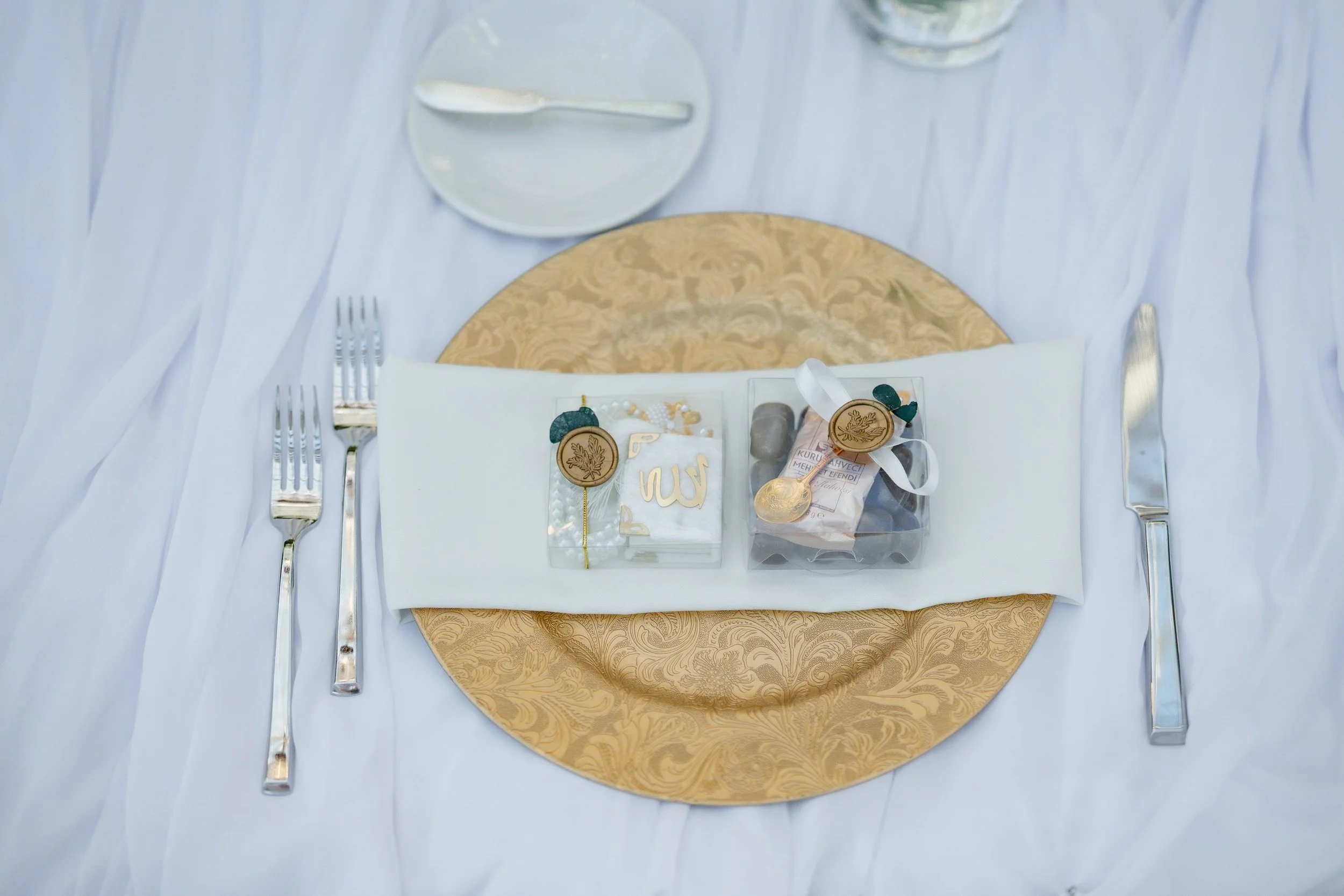 Wedding table setting with a gold decorative placemat, silverware, a white napkin, and a gift box with jewelry and a tag on a white tablecloth.