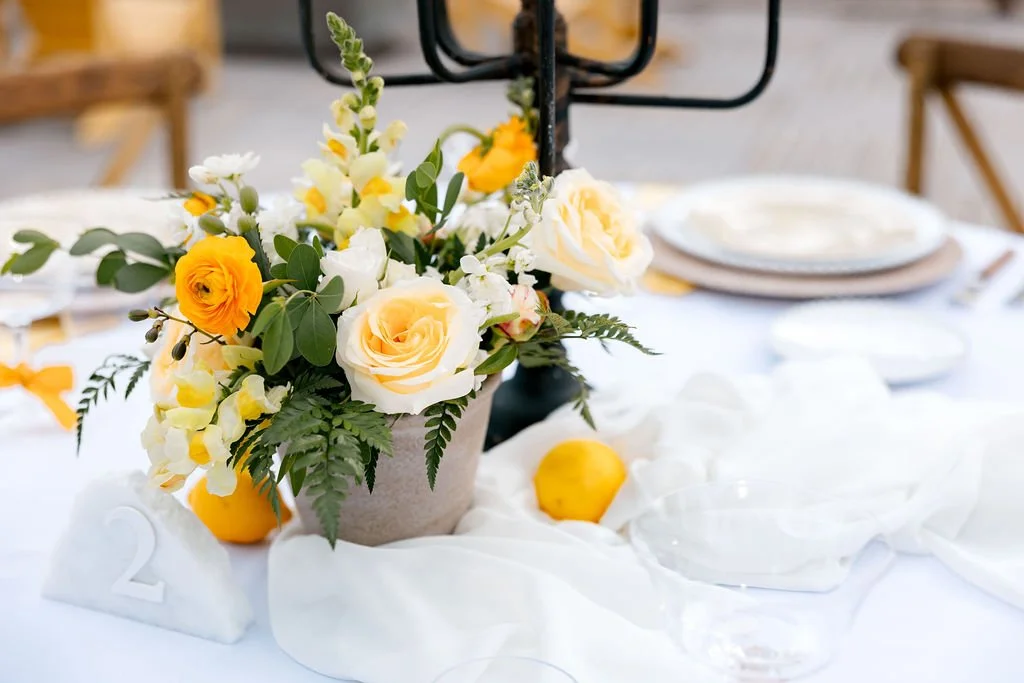 A floral centerpiece with white and yellow roses, greenery, and lemons on a white tablecloth at an outdoor event.