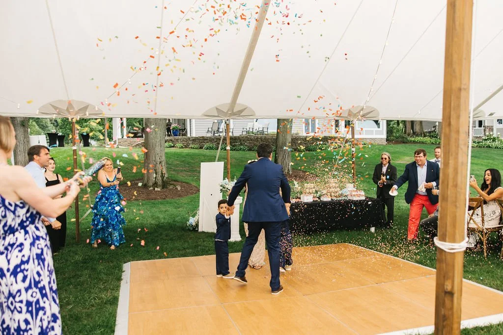 People celebrating outdoors under a tent with confetti and sparklers, including a man spinning a child, with others watching and taking pictures.