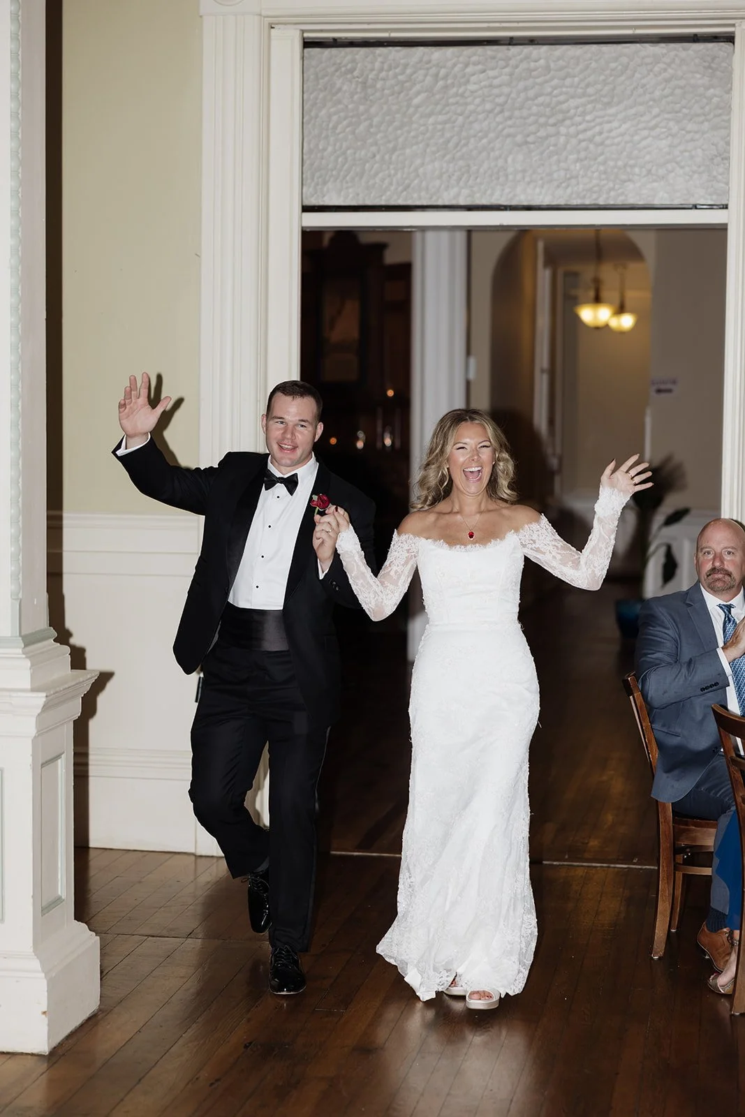 A wedding celebration with a bride and groom walking into the room, smiling and waving, while a guest sits at a table nearby.
