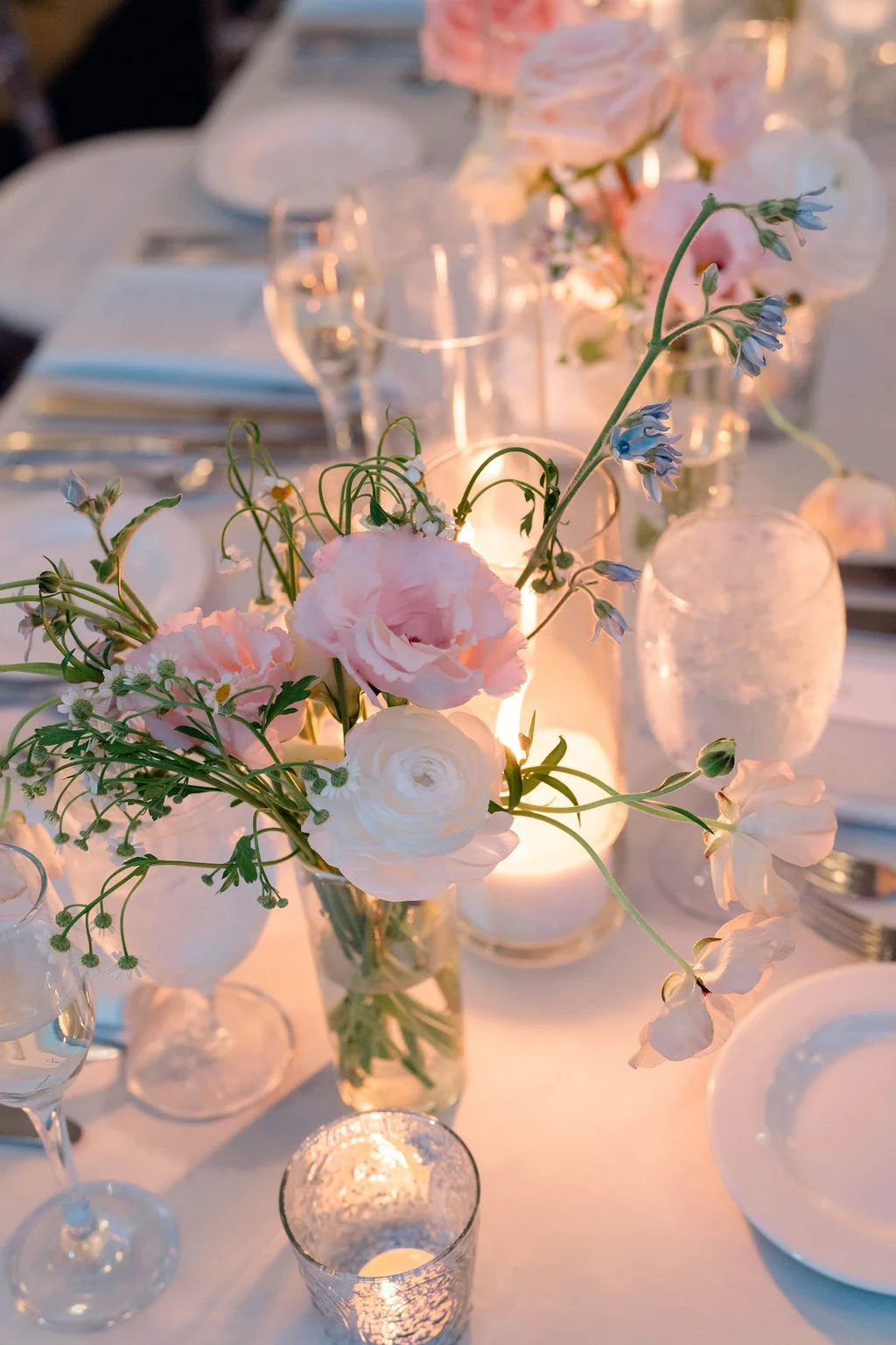 Close-up of a table centerpiece with soft pink and white flowers, candles, and glassware at an elegant event.