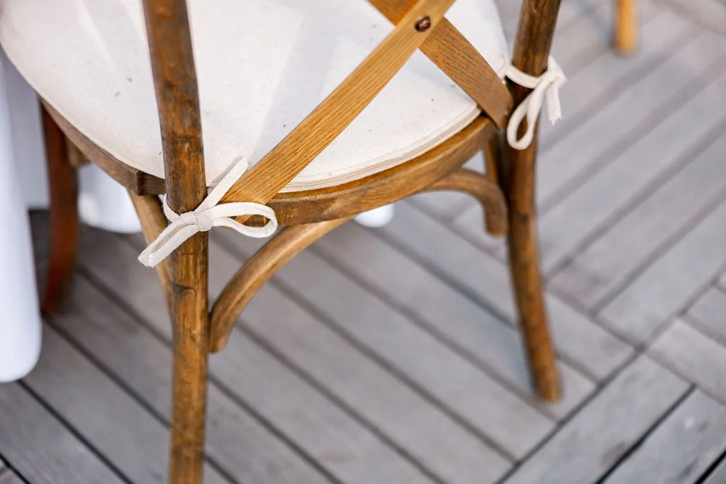 A wooden chair with a light-colored seat and tied white ribbons on the back and side, placed on a wooden deck floor.