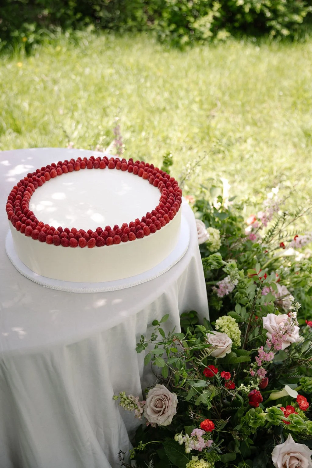 A white cake with red berries on top, placed on a white tablecloth and surrounded by colorful flowers.