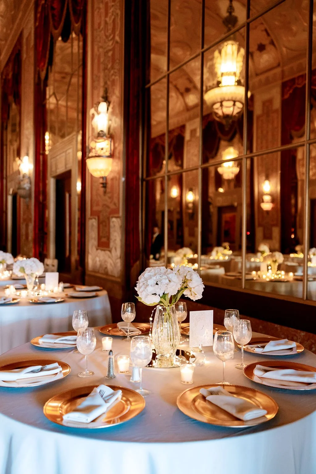 Elegant banquet table with white tablecloth, gold charger plates, white napkins, wine glasses, candles, and a floral centerpiece with white flowers in a clear vase. Warm lighting and ornate wall decor reflect in a mirror.