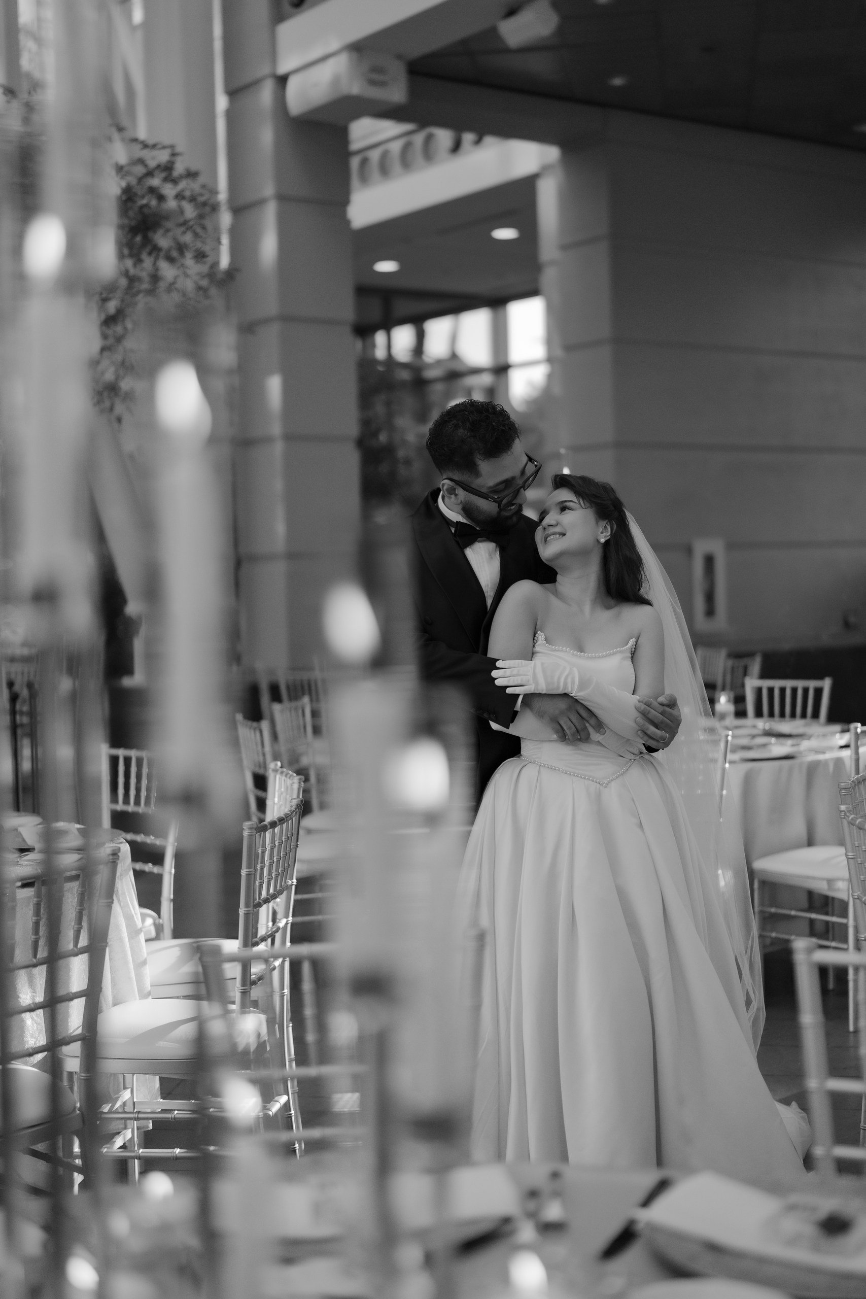 A black and white photo of a bride and groom dancing at a wedding reception indoors. The groom is wearing glasses and a tuxedo, and the bride is wearing a strapless wedding gown and veil. They are gazing into each other's eyes, smiling, with tables a