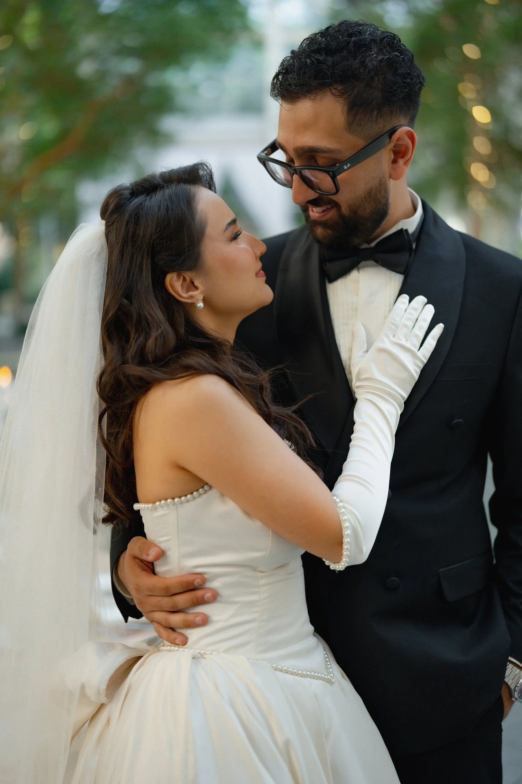 A bride and groom dancing closely at their wedding, with the bride wearing a white dress and pearl accessories, and the groom in a black tuxedo and glasses.