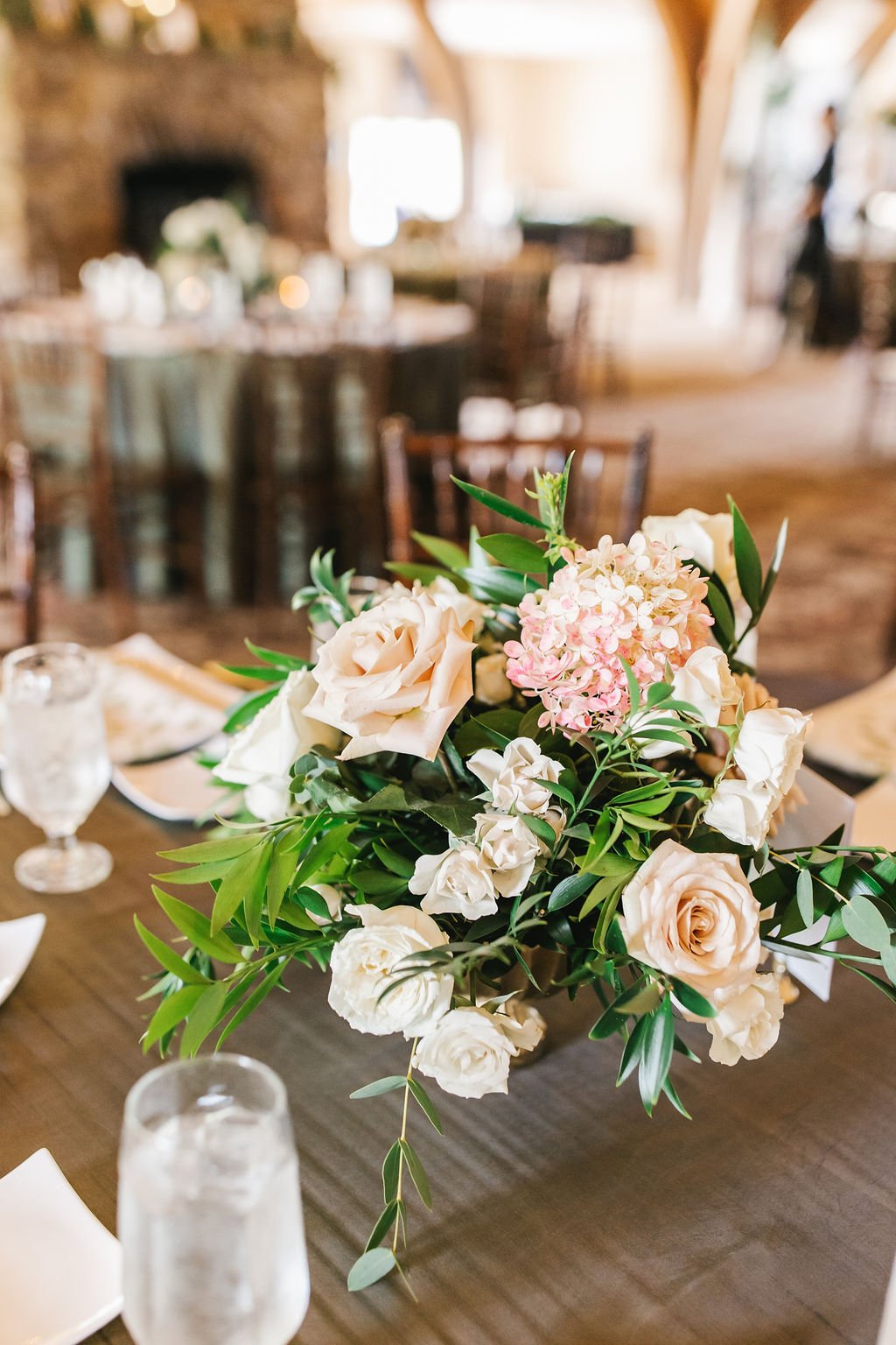 A floral centerpiece with peach and white roses, pink hydrangeas, and greenery on a wooden table in a rustic, well-lit venue.