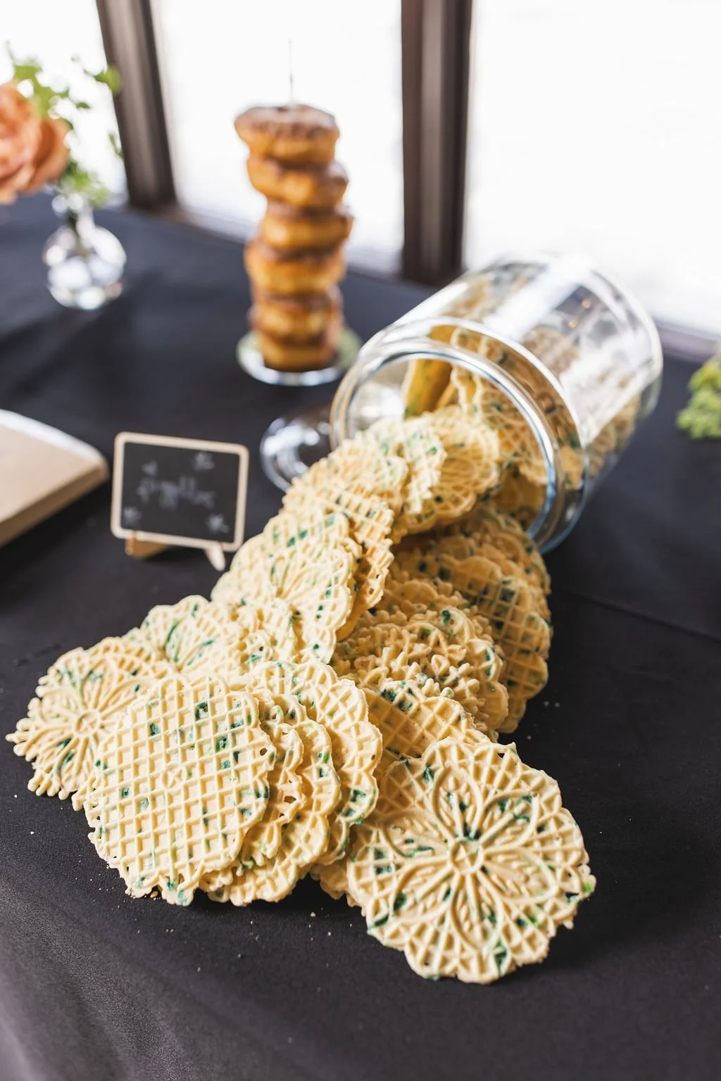 A black tablecloth covered display with decorative waffle-shaped cookies spilling from a glass jar, with a stack of cookies in the background and a small blackboard sign nearby.