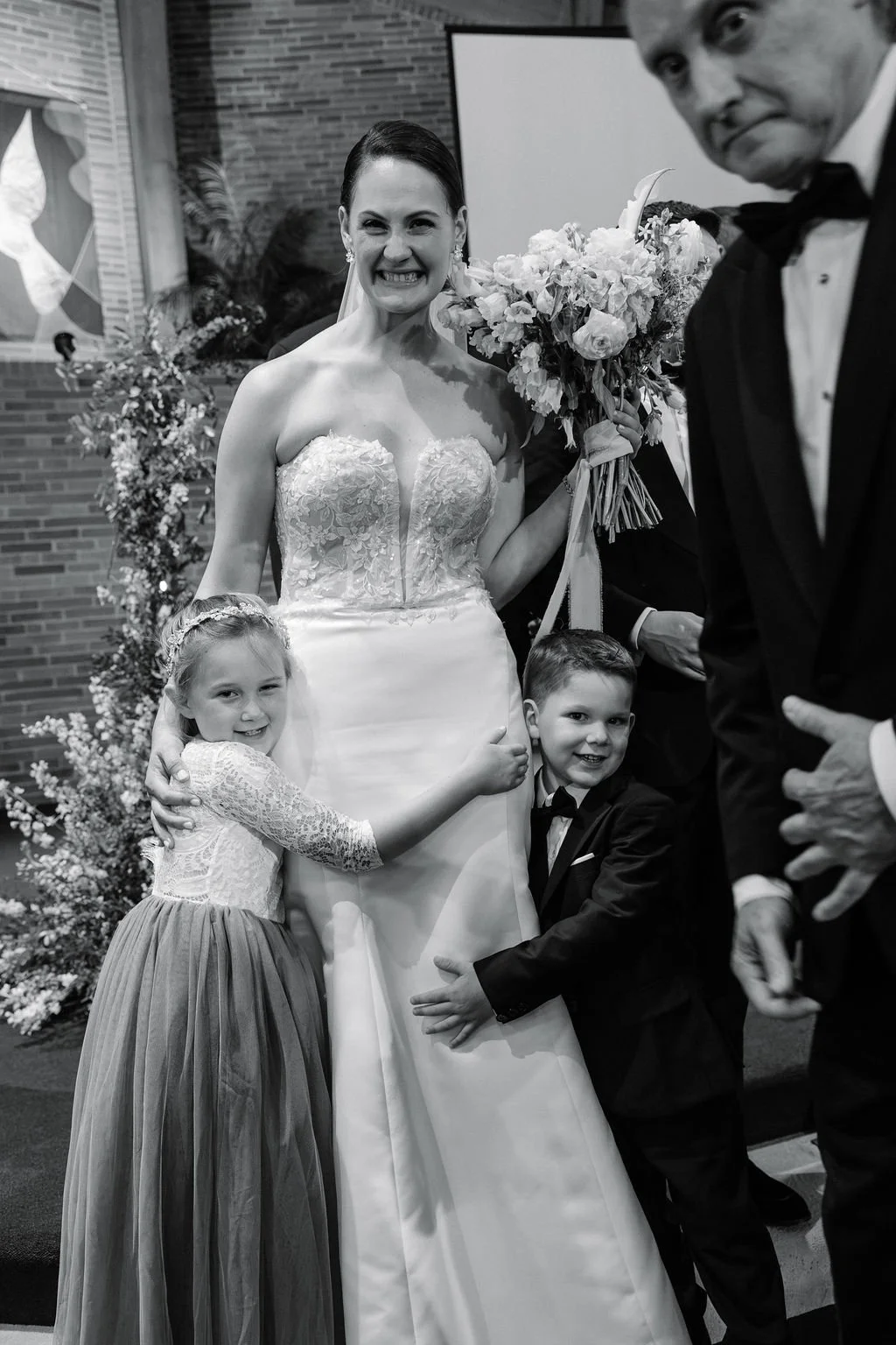 A bride in a wedding dress holding a bouquet, smiling, with two children hugging her, a girl and a boy, in formal attire, at a wedding ceremony.