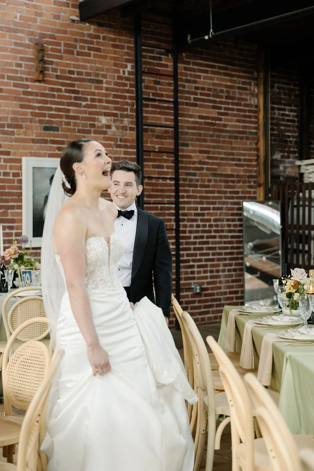 A bride and groom at their wedding reception, with the bride smiling and the groom looking at her, in a brick-walled venue decorated with flowers and table settings.