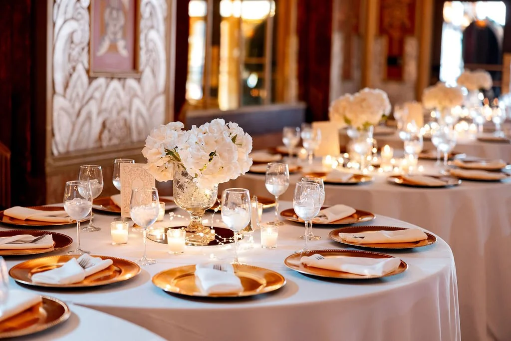 Elegant banquet table decorated with white flowers in a glass vase, surrounded by candles, with gold chargers, white napkins, and water glasses, set in a warmly lit rustic venue.