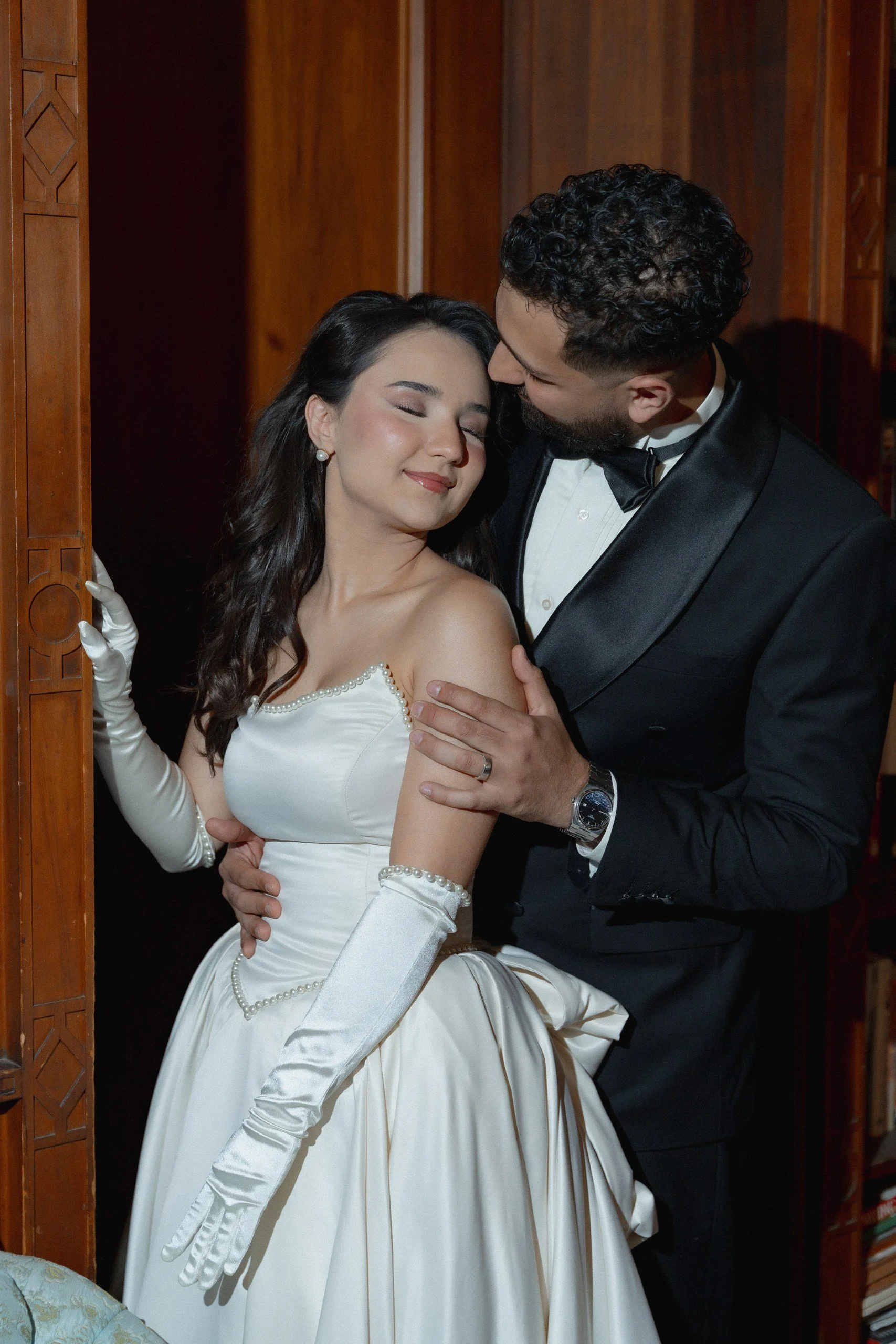 A bride and groom in wedding attire share an intimate moment, with the groom kissing the bride's forehead as she closes her eyes, standing in front of a wooden wall.