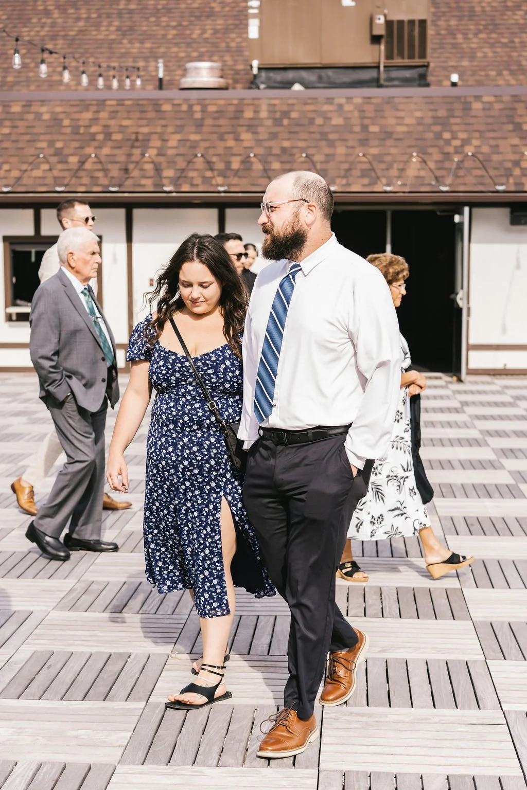 People walking outdoors on a wooden deck in sunlight, including a woman in a blue floral dress and a man in dress shirt and tie, with other individuals in the background.