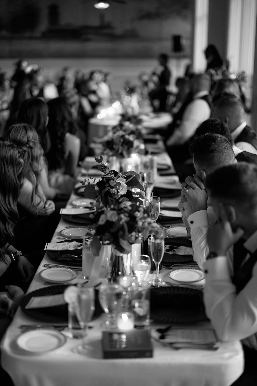 Black and white photo of a formal dinner gathering with people seated at a long table decorated with floral arrangements, candles, and place settings, with some people appearing to pray or reflect.