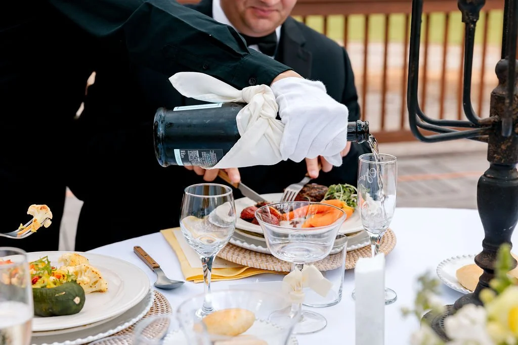 Waiter pouring champagne into a glass at an outdoor restaurant table with food, wine glasses, and decorative elements.