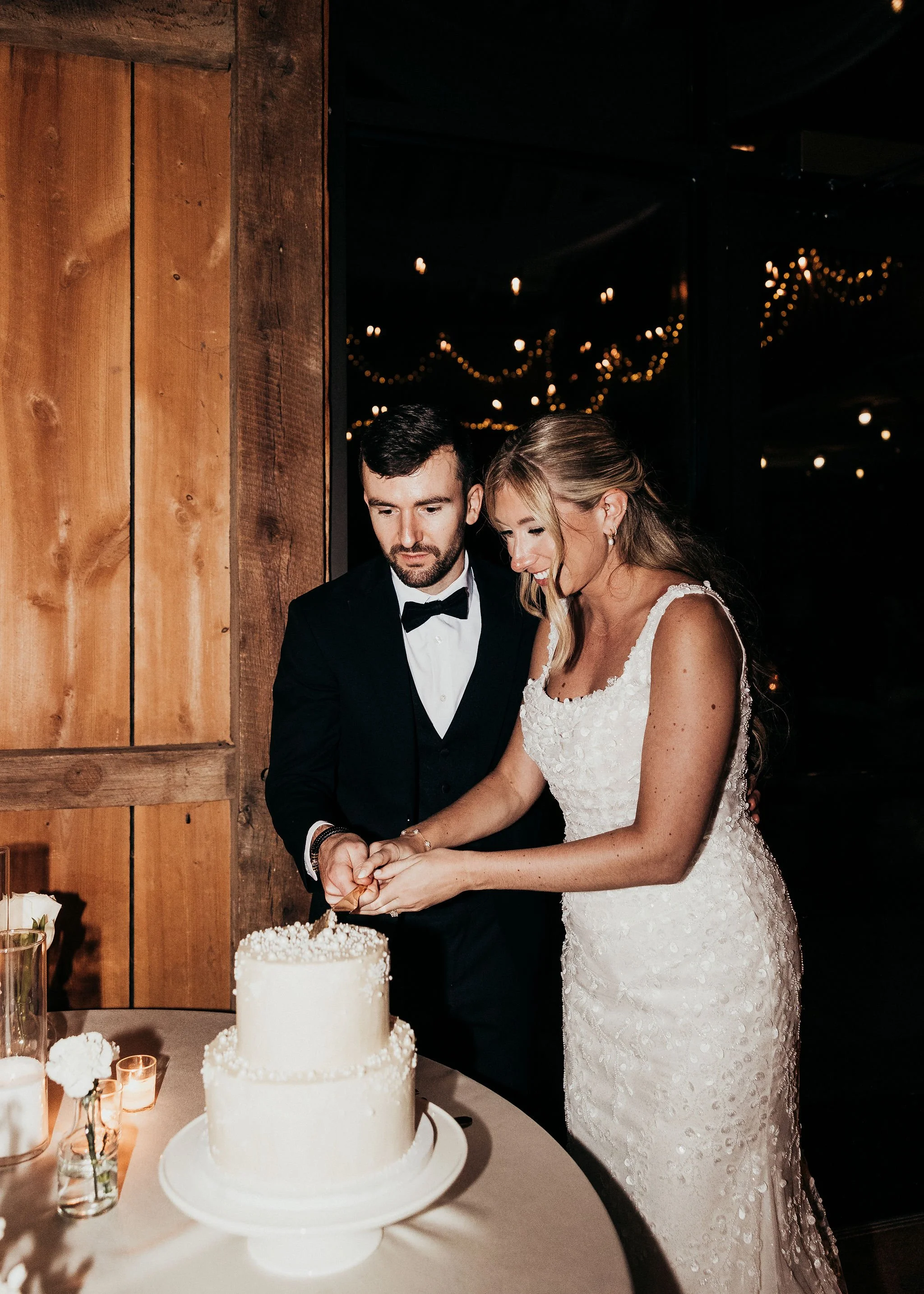 A bride and groom cut a wedding cake together at their wedding reception, with the bride wearing a white lace gown and the groom in a black tuxedo.
