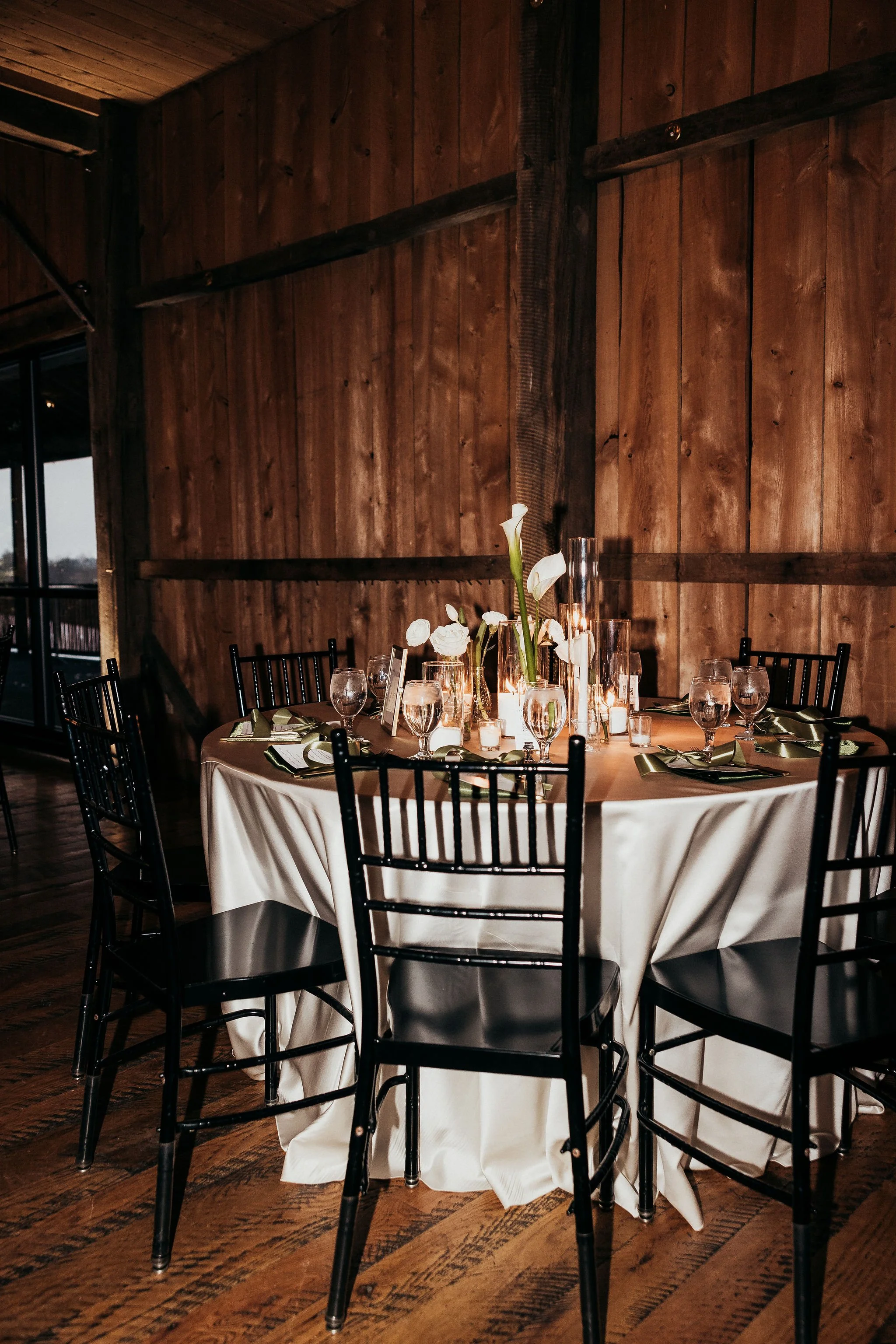 A round table covered with a white tablecloth, decorated with a tall white floral centerpiece, surrounded by black chairs, and set with glasses, plates, and green napkins. The background features a wooden wall and large windows.