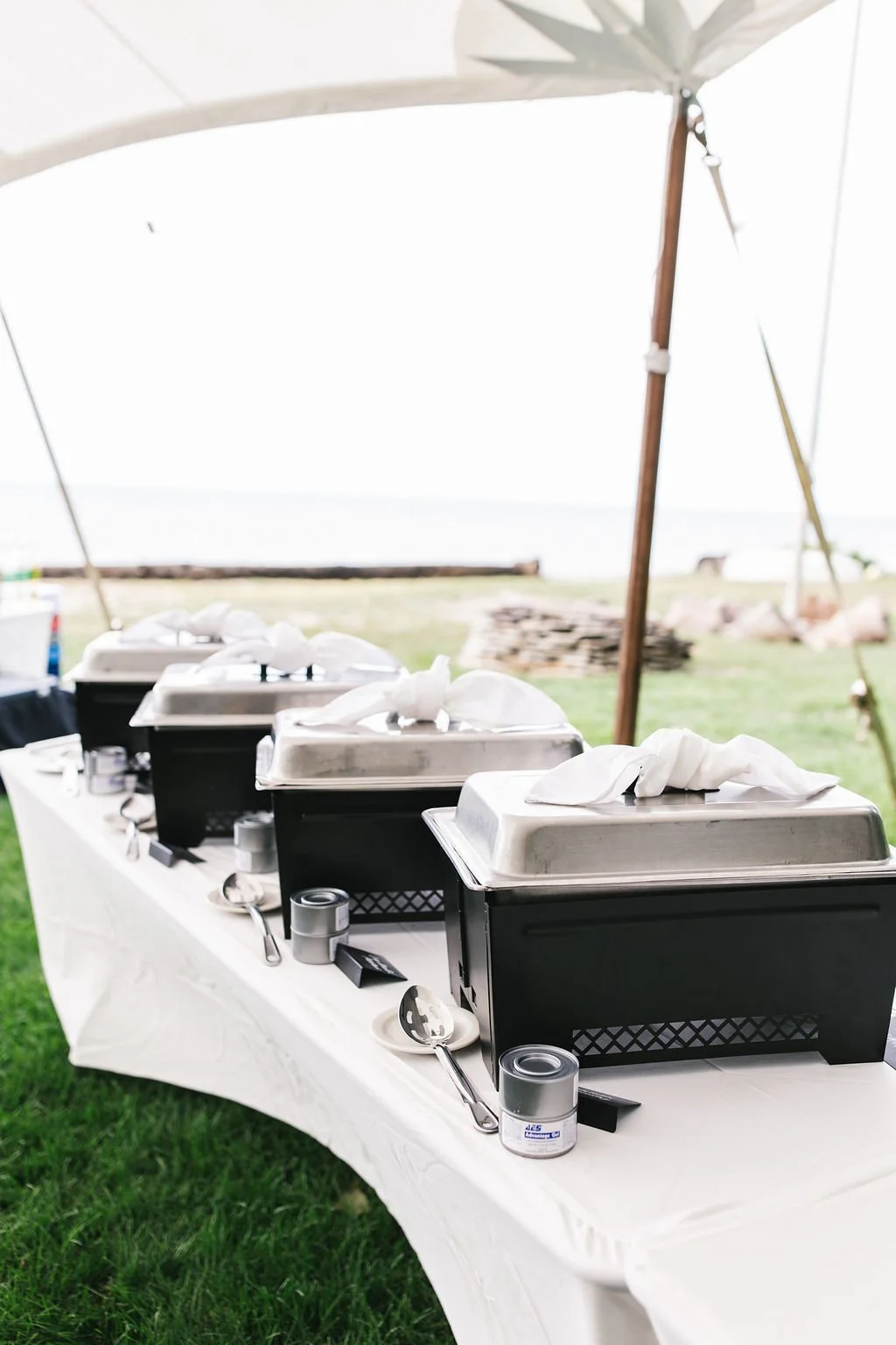 Chafing dishes on a buffet table set up under a canopy at an outdoor event, with cutlery and cans of food nearby, and a grassy area in the background.