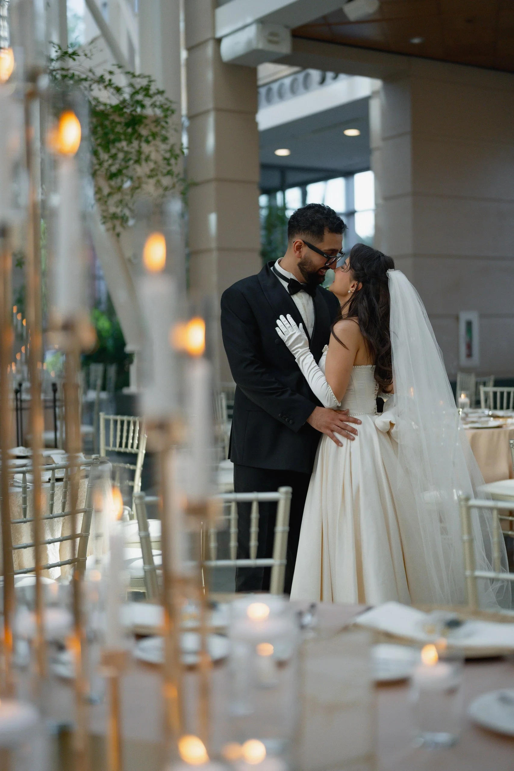 A bride and groom sharing a moment at their wedding reception indoors, with decorated tables and candle holders in the foreground.