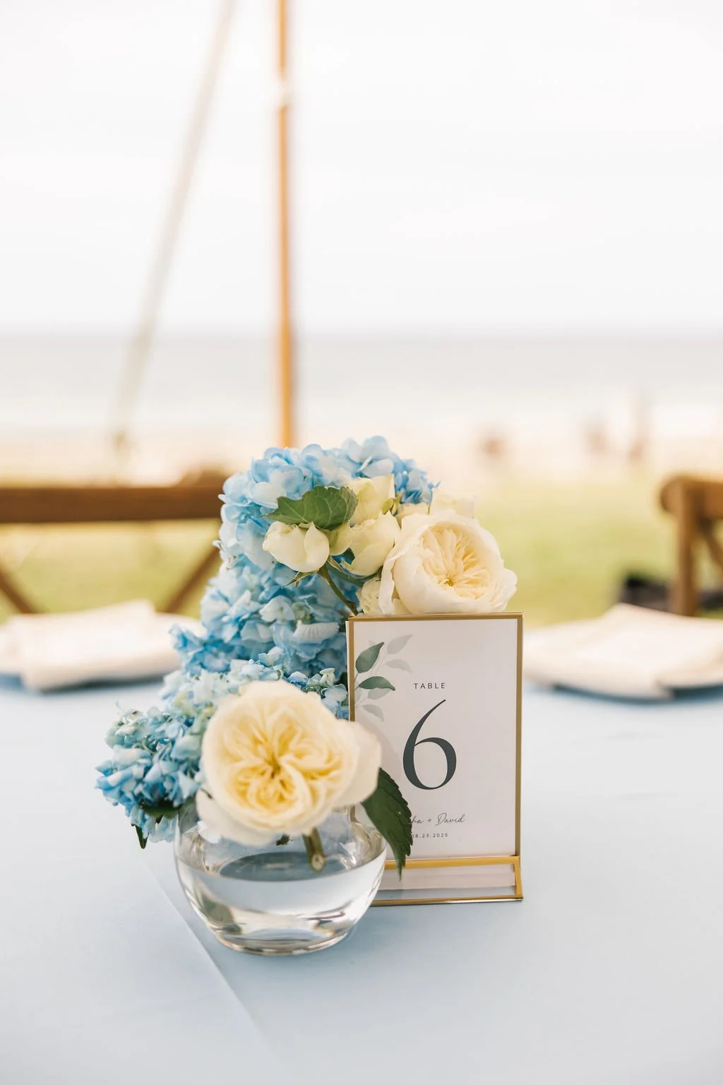 A floral centerpiece with white roses and blue hydrangeas on a table at an event, with place cards and table number 6 visible.