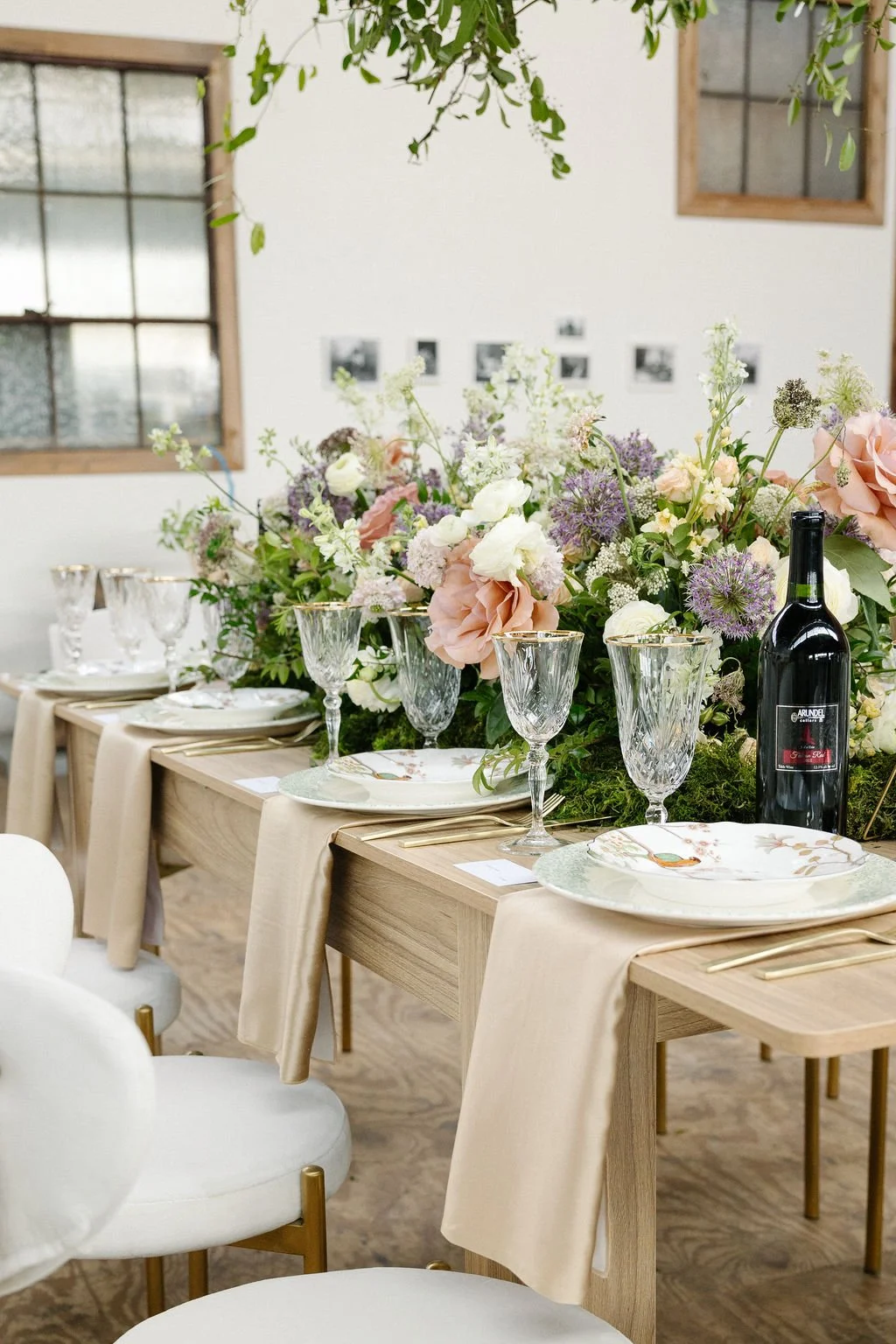 A decorated dining table with elegant glassware, floral arrangements, and a bottle of wine, set for a special occasion in a bright room with windows.