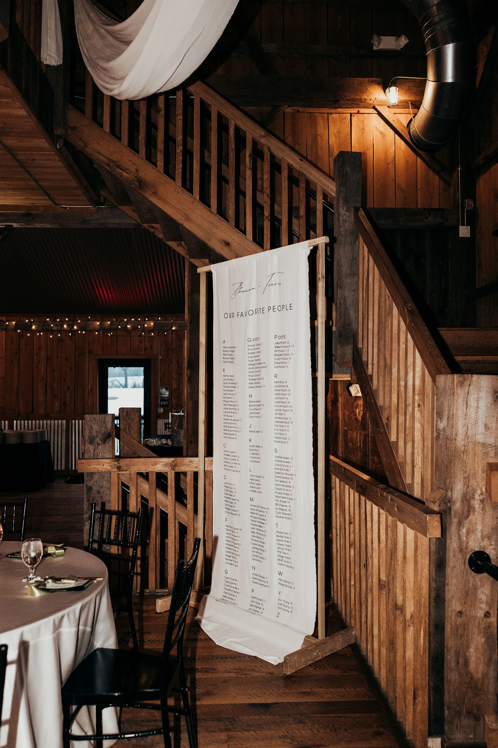 Indoor view of a rustic wooden banquet hall with a staircase, a hanging fabric banner, a seating area with round tables covered in white tablecloths, black chairs, and a large vertical seating chart with names listed, decorated with string lights.