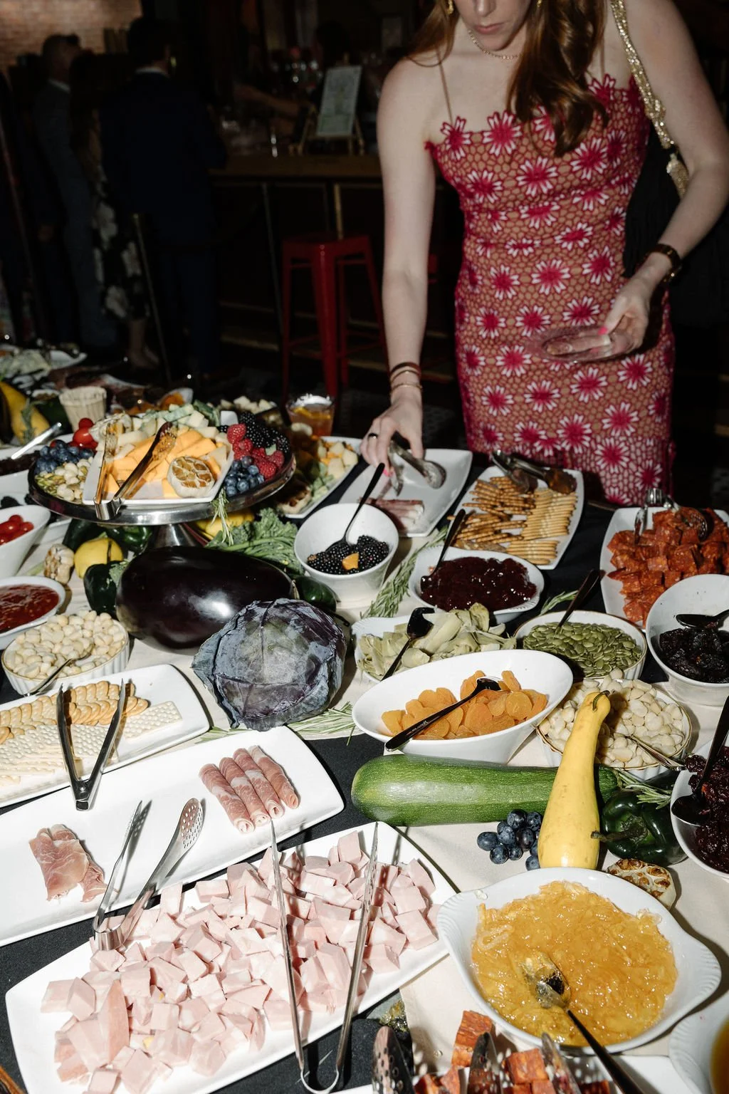 A woman in a red floral dress selecting food from a buffet table filled with fruits, cheeses, meats, and jams at a social gathering.
