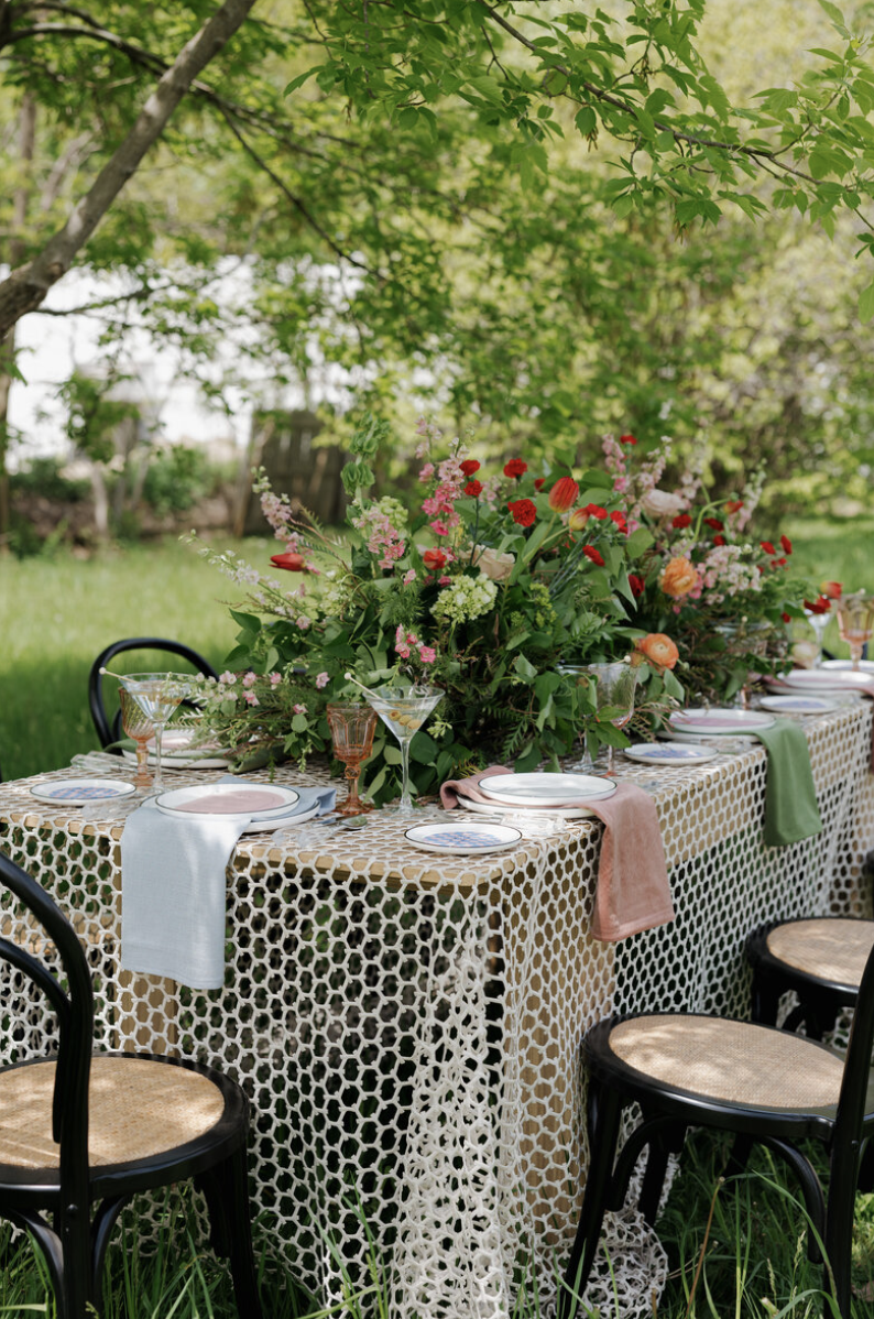 Outdoor dining table set with a large floral centerpiece, surrounded by chairs with woven seats, under a leafy tree in a lush garden.