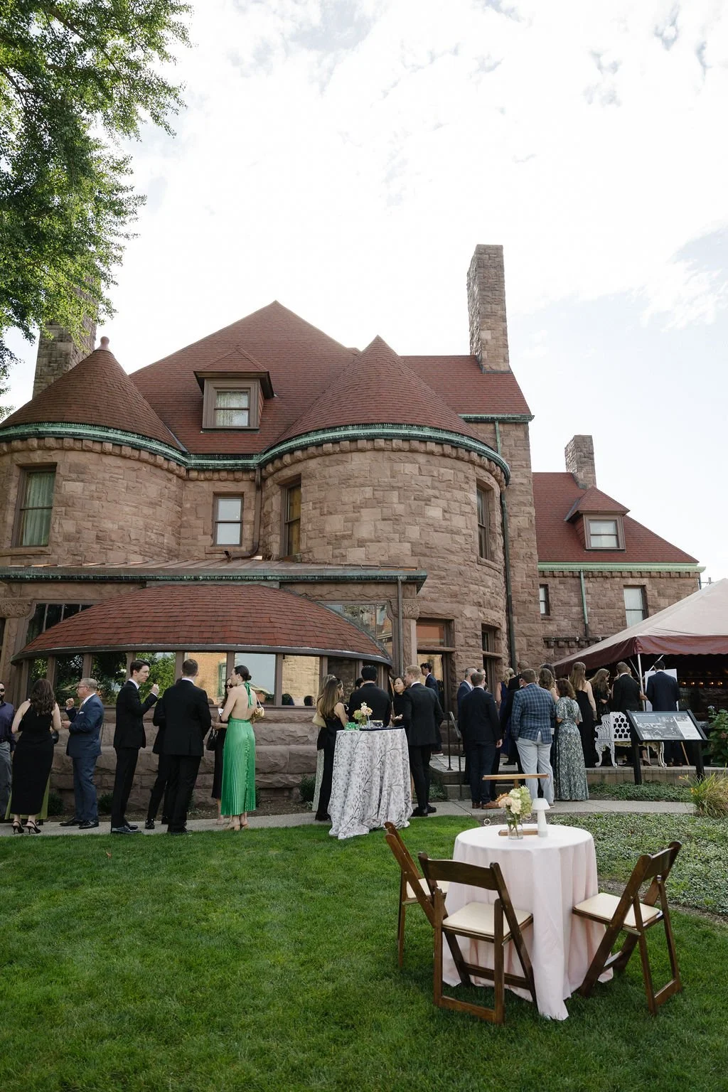 Guests gathered outside a large stone mansion for a social event, standing near a table with a white tablecloth, with additional tables and chairs set up on the lawn.