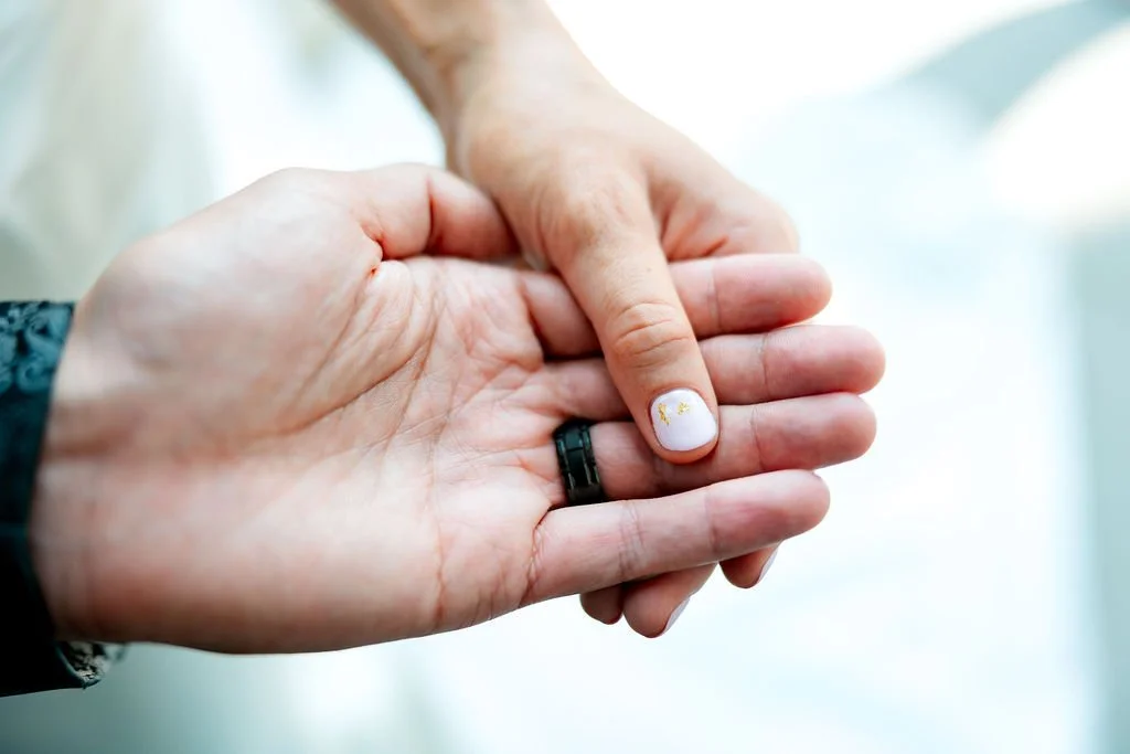 Close-up of two hands holding each other, one hand with a wedding ring and painted fingernails, the other hand with a dark bracelet, against a blurred light background.