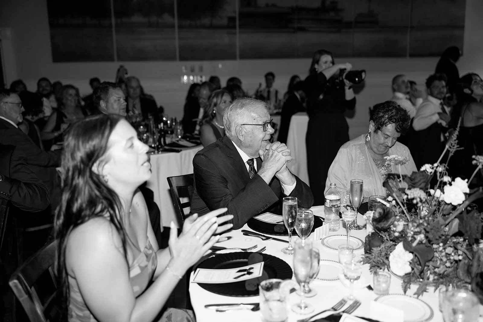 People seated at a formal dinner event, some with drinks, with a floral centerpiece, and someone taking photographs in the background.