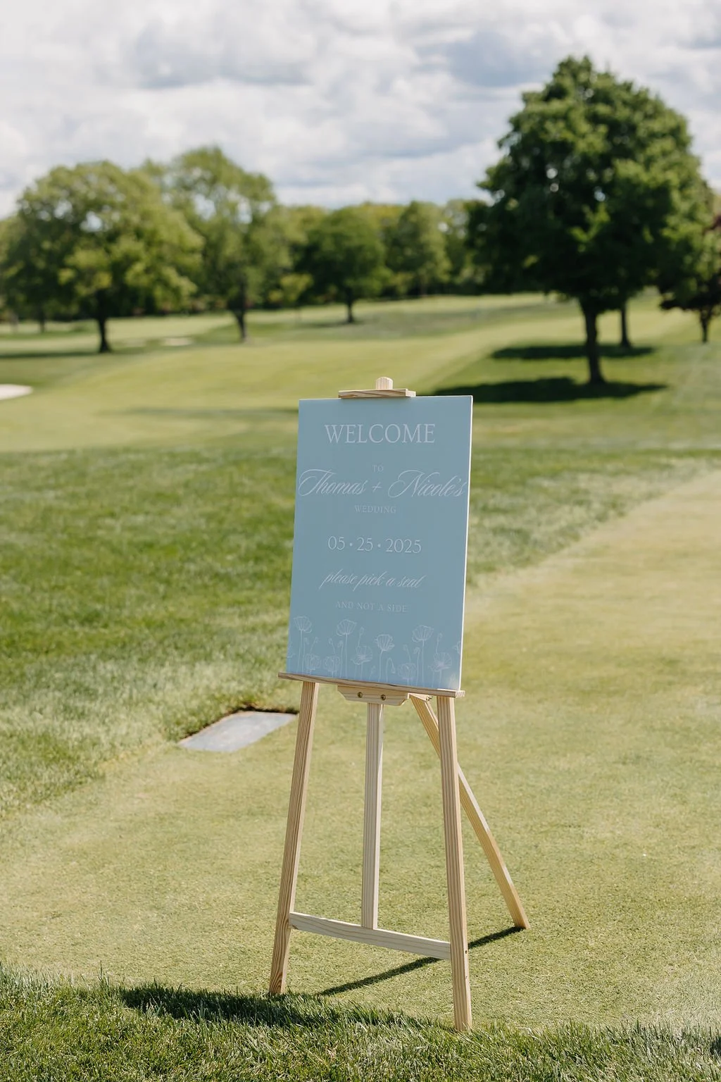 A wedding sign on a wooden easel in a golf course, welcoming guests to Thomas and Nicole's wedding on May 25, 2025, with a background of green grass, trees, and partly cloudy sky.