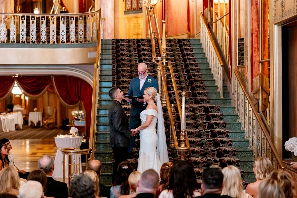 Bride and groom holding hands during their wedding ceremony on a staircase, with officiant standing behind them and guests seated watching.