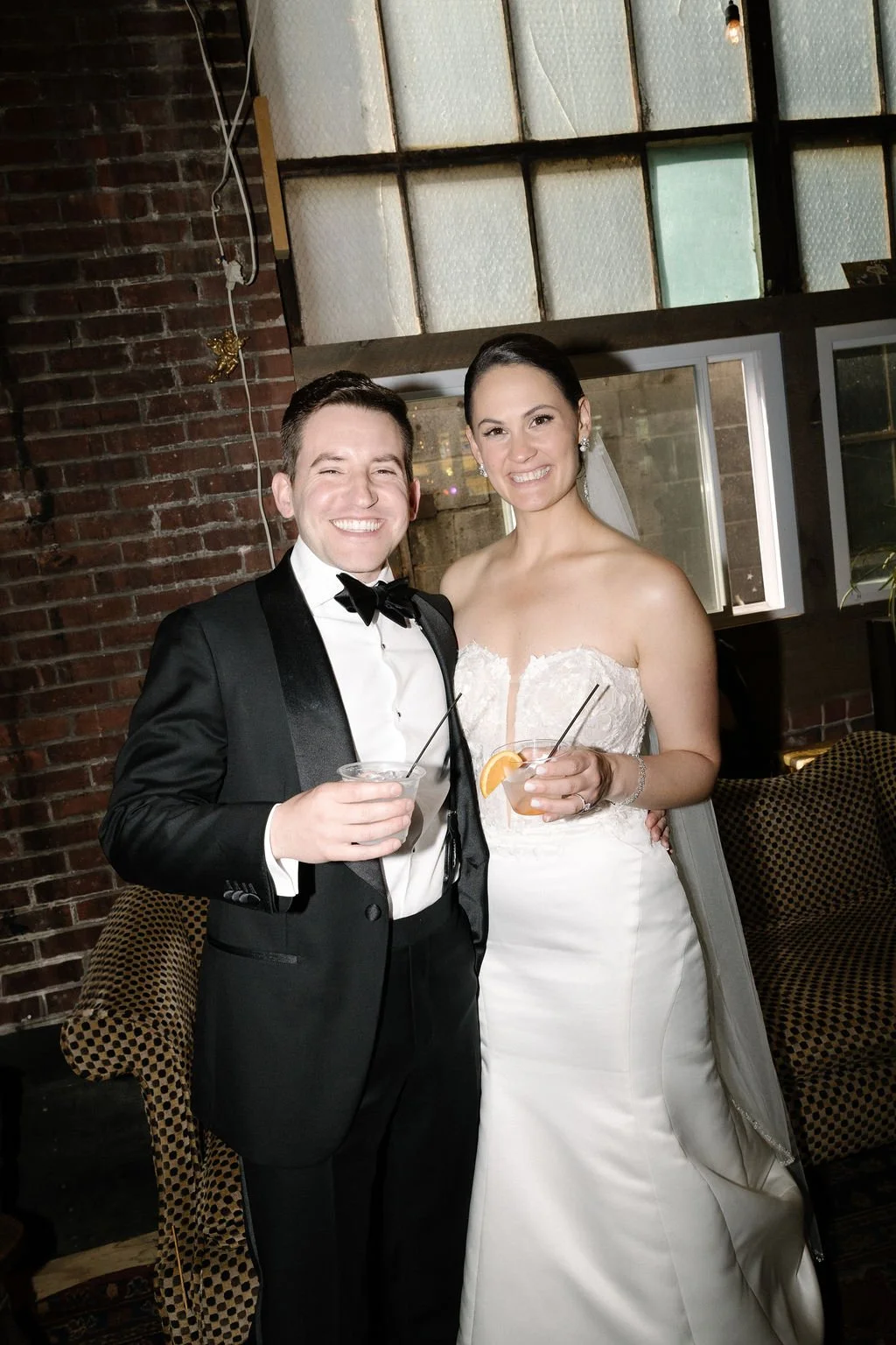 A newlywed couple smiling and holding drinks in celebration during their wedding reception. The groom is wearing a black tuxedo with a bow tie, and the bride is in a strapless white wedding gown with a veil.