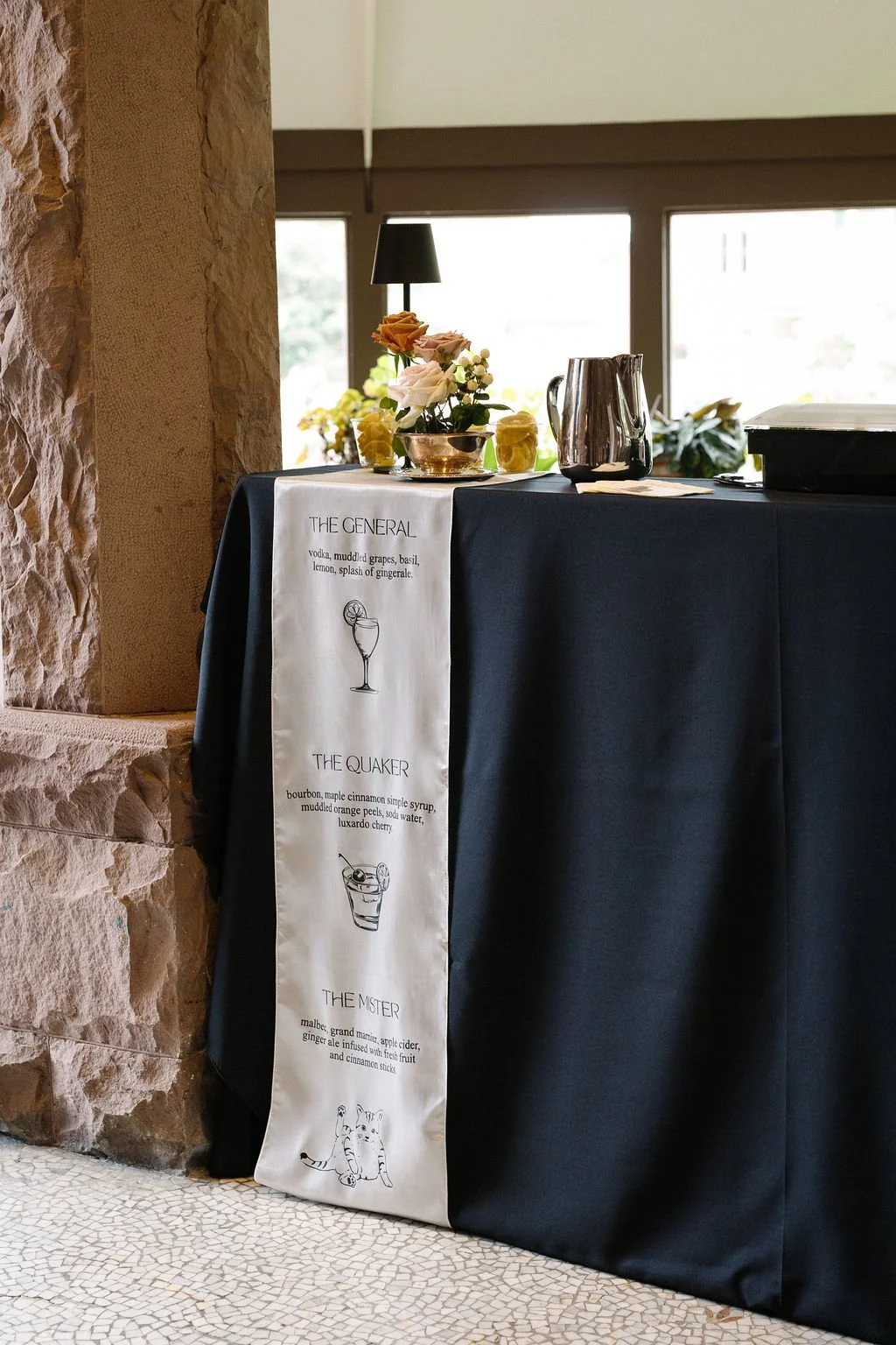 A table covered with a dark tablecloth and a white step-and-repeat banner featuring drink recipes and illustrations. The table holds a small flower arrangement, a black lamp, and metal pitchers, with sunlight coming through windows in the background.