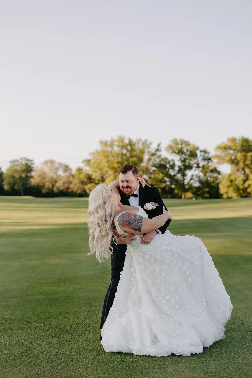A newlywed couple dancing on a grassy field during sunset, with the groom holding the bride and smiling, trees in the background.