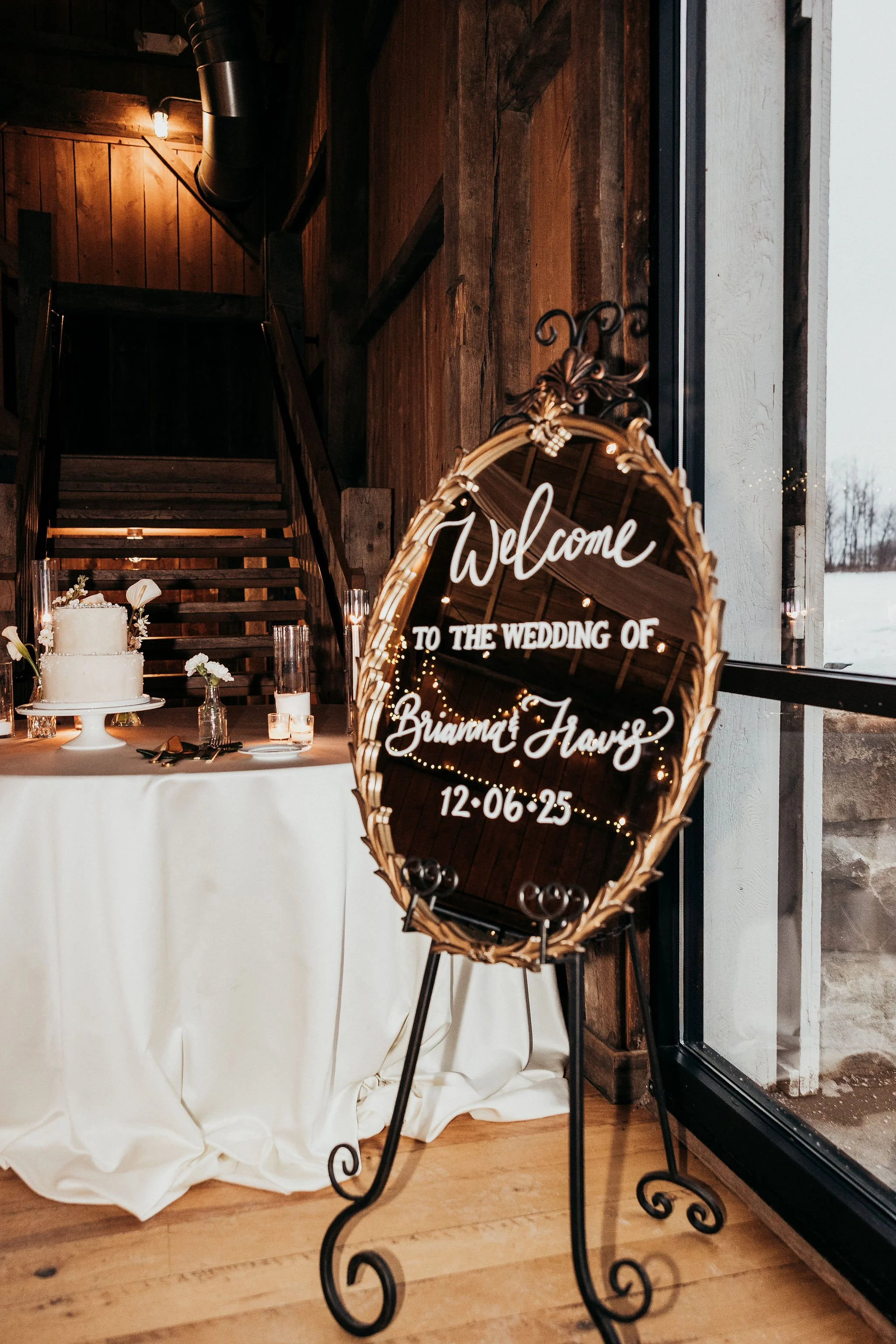A decorative mirror with a lit LED sign that reads 'Welcome to the wedding of Brianna & Travis 12-06-25', placed in front of a table with a white tablecloth and wedding decor, inside a rustic venue with wooden walls and staircase.