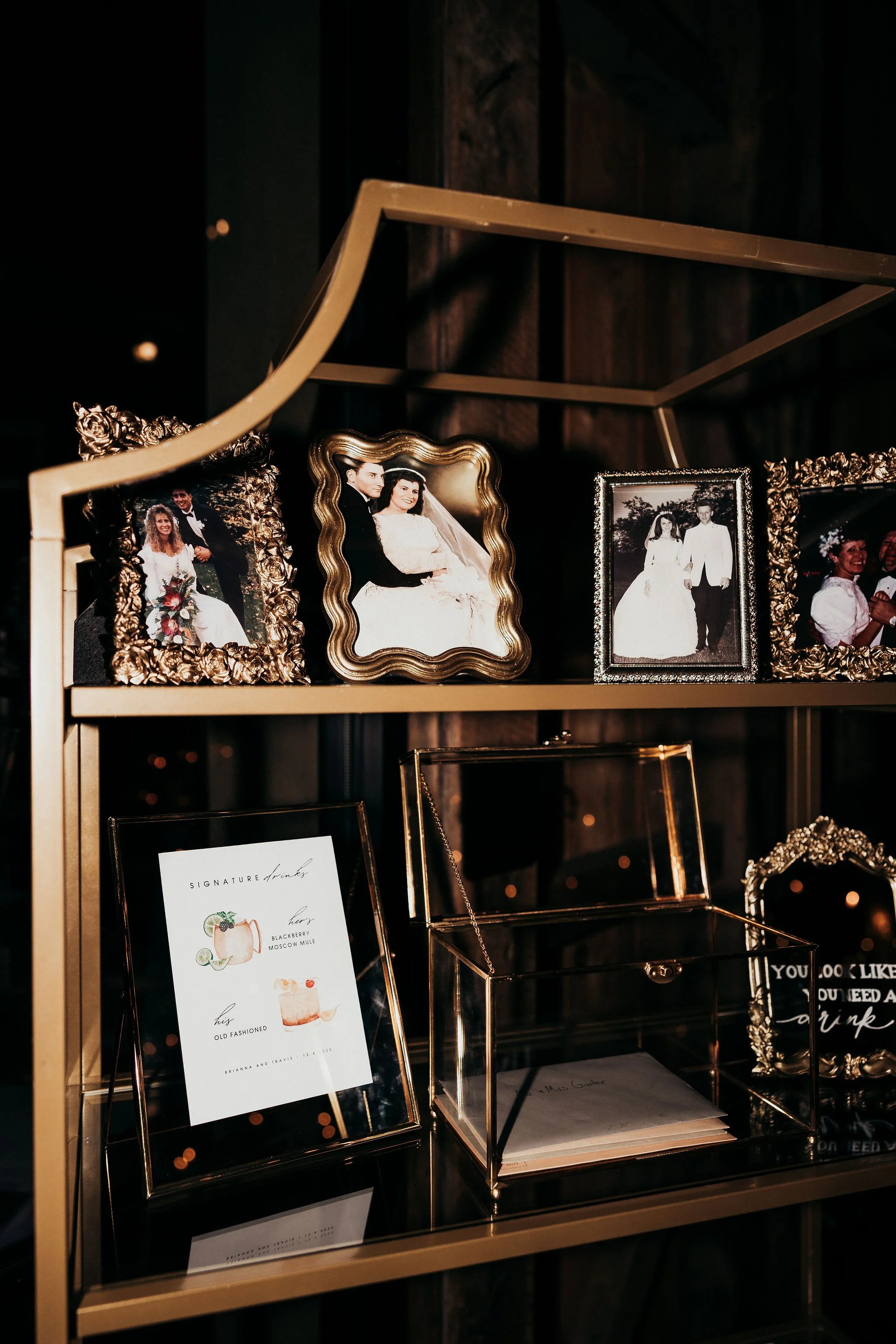 Display of framed photographs of a wedding couple, several decorative picture frames, a menu with cocktail list, and glass display cases on a gold-colored shelf.