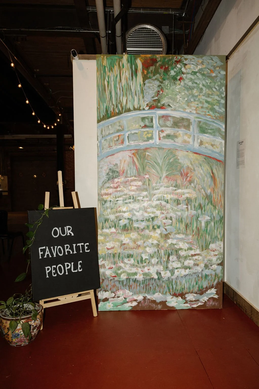 A colorful painting of a garden scene with a small arched bridge over water, lily pads and flowers, displayed indoors next to a blackboard sign that reads "OUR FAVORITE PEOPLE".